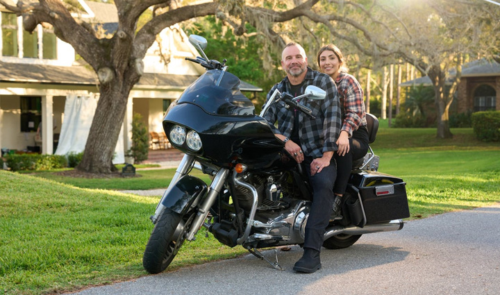 Wounded Warrior Bill Geiger and his daughter sitting on motorcycle parked on a residential street, surrounded by greenery with sunlight filtering through the branches.