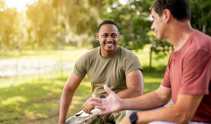 Warriors Tyshawn Jenkins and Michael Albaugh sit and talk outdoors in a park-like setting with trees and sunlight in the background.