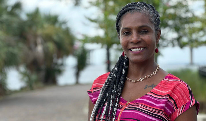 Wounded warrior Tonya Oxendine wears a brightly colored blouse with pink, purple, and yellow with her long braid draped over her shoulder in front of a background of blue sky and green palm trees. 