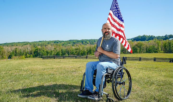 Warrior Paul Guzman sits in a wheelchair outdoors in a grassy field holding an American flag, with rolling hills, trees, and a blue sky in the background.