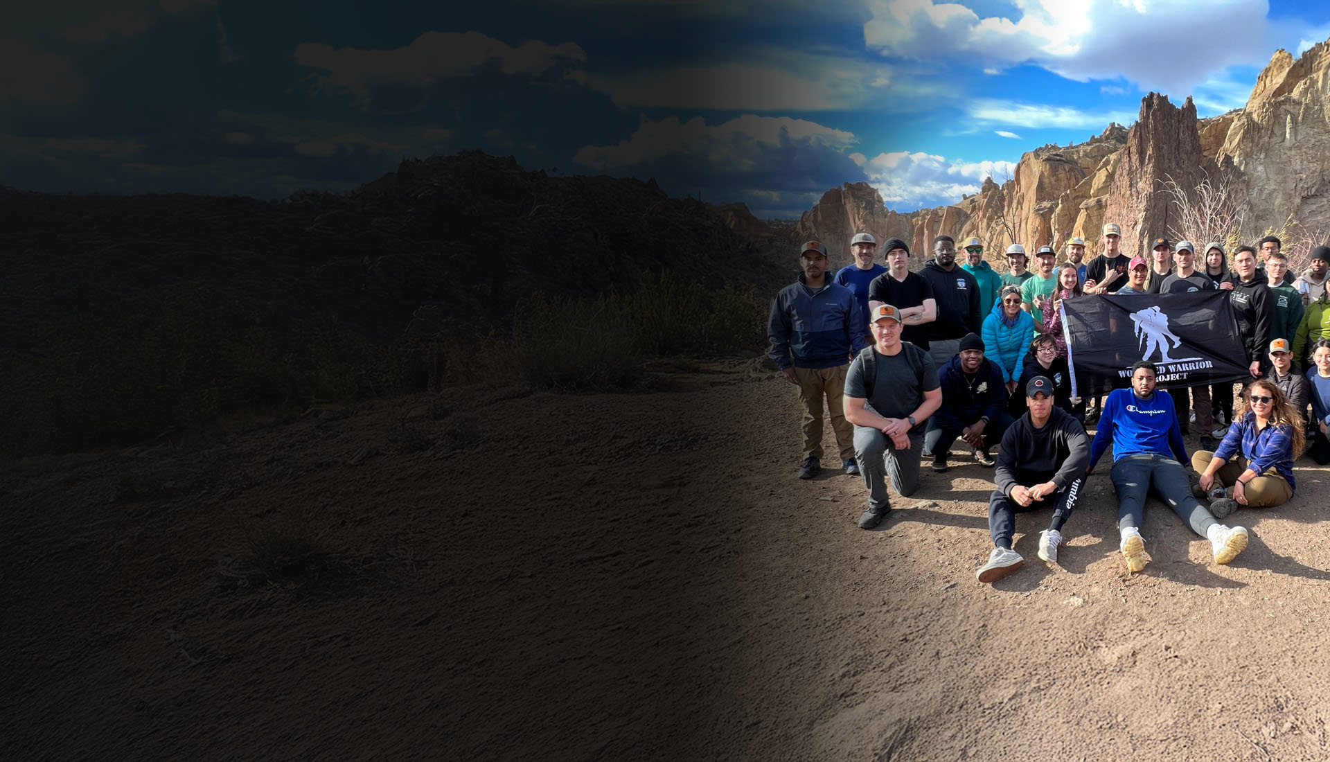 A group of wounded warriors pose for a group photo in front of mountains and smile while holding a black flag with a white WWP logo.