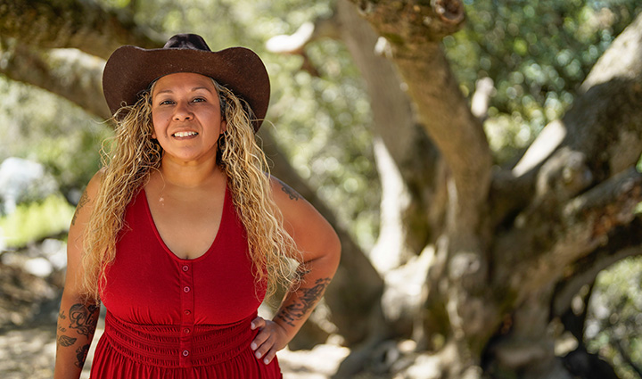 Wounded warrior Ysable Carona stands in front of a large tree while smiling at the camera with her left hand on her hip.  