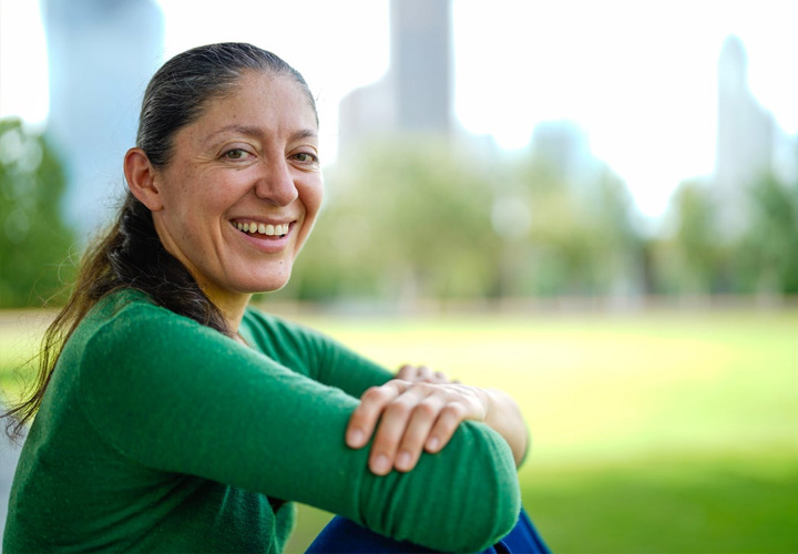 Wounded warrior Ruth Corrigeux sits with her arms crossed while smiling at the camera. She’s out on a grassy field with a skyline in the background.