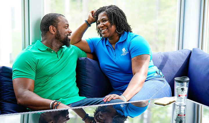 Warrior Taniki Richard, wears a blue polo, sits on her couch, talking with her husband, Brandon, who is wearing a green polo.