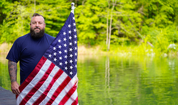 Wounded warrior Matthew Templeton holding an American flag near water with trees in the background.