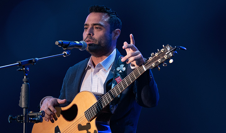 Wounded warrior Sal Gonzalez stands on stage speaking into a microphone while holding his guitar.