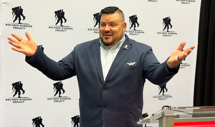 Veteran Jake Norotsky speaks on stage in a blue suit jacket in front of a white backdrop with WWP logos.