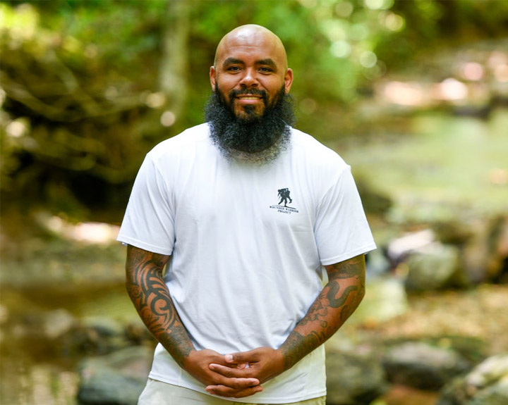 Wounded warrior Eric Delion stands in front of a stony brook surrounded by trees while clasping his hands and smiling at the camera.