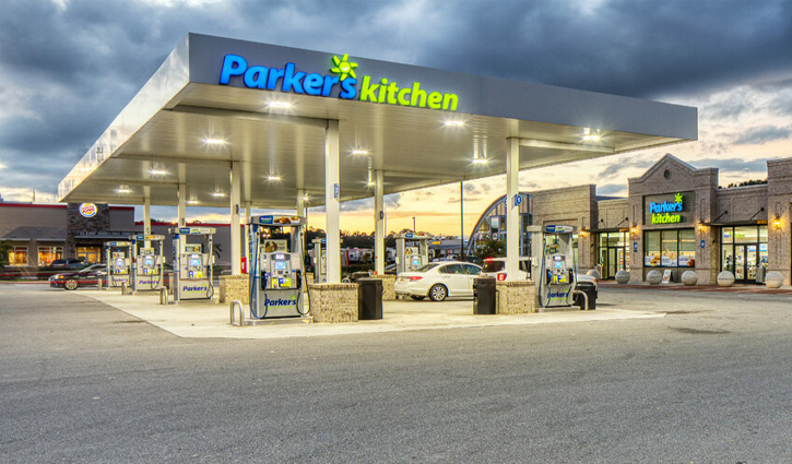 A Parker's Kitchen convenience store with gas pumps in front of it and its blue and green signs lit up with an evening sky in the background.