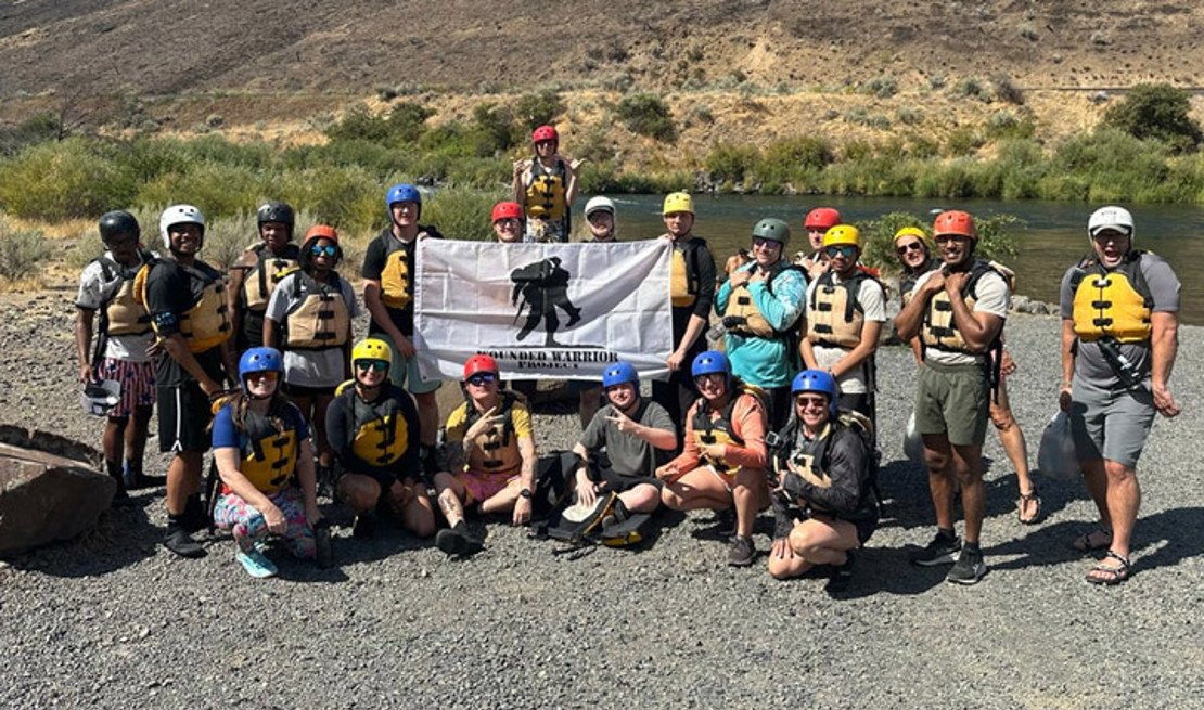 A group of active-duty service members pose together holding a Wounded Warrior Project flag after whitewater rafting.