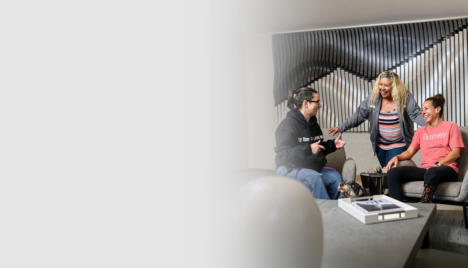Three warriors sit and converse in a modern lounge, with a small dog nearby and a textured wall behind them.