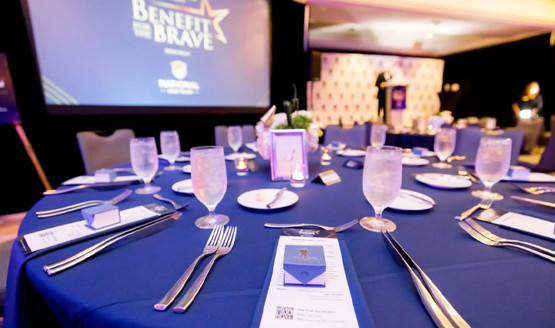 Benefit for the Brave table with blue cloth, menus, glasses, and utensils. There is a presentation screen in the background.