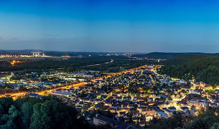 Panoramic view of Landstuhl at dusk with glowing town lights, forested hills, and a clear sky fading from dark to light blue. 