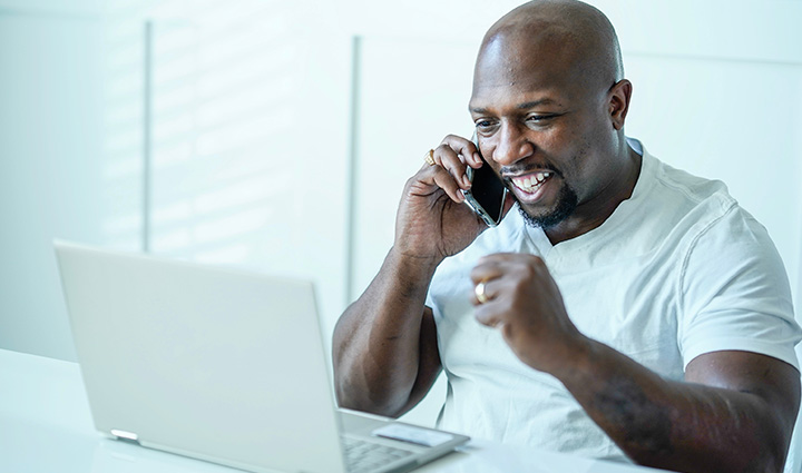 Warrior Melvin Gatewood smiles and raises his left arm with excitement, looking at his computer with a cellphone up to his right ear.