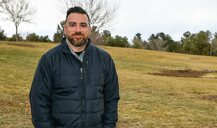 Wounded warrior Arnulfo Dauto stands outside with leafless trees in the background. He wears a jacket while smiling at the camera. 