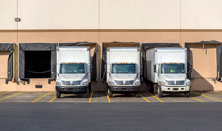 Three semi-trucks with box trailers parked in row at a warehouse.