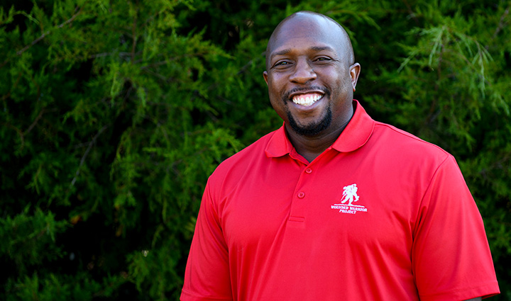 Wounded warrior Melvin Gatewood stands in front of a large shrub while wearing a red WWP polo shirt and smiling at the camera. 