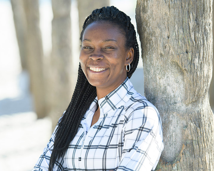 Wounded Warrior Danielle Green leans against a tree at the beach, arms crossed and smiling.