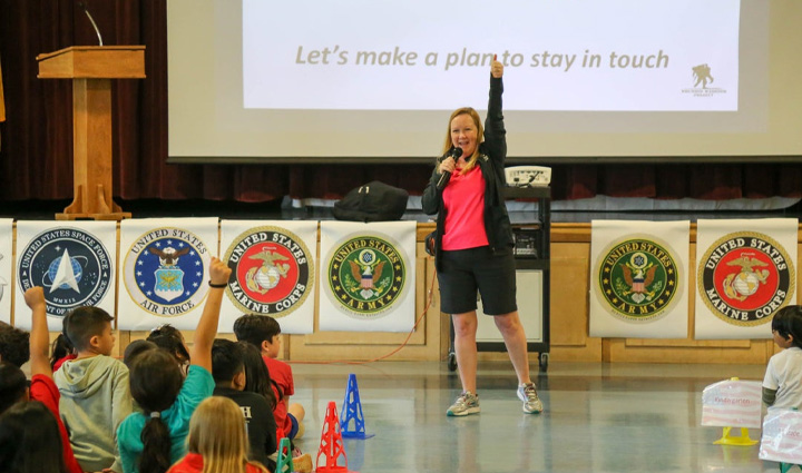 Jennifer Jenkins raises her right arm in the air while smiling and engaging a school of children to do the same.