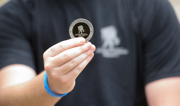 Close-up of a warrior holding a WWP challenge coin. The commemorative coin features the WWP logo and the phrase “The Greatest Casualty Is Being Forgotten”. 
