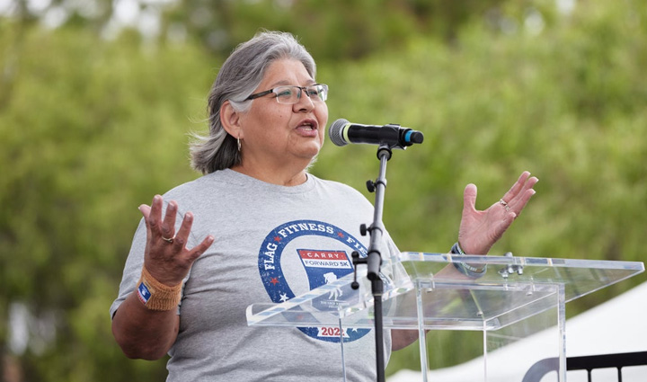 Gold Star Mother Mary Tallouzi wears a grey shirt and stands at a podium with trees in the background as she speaks at a WWP Carry Forward 5K event.
