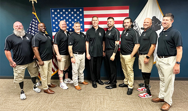 A group of nine warriors wearing black polo shirts stand in front of the American flag while smiling at the camera.