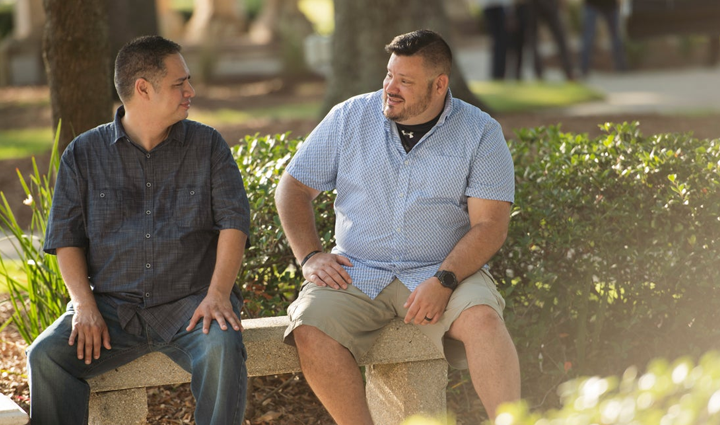 Warrior Jake Norotsky, wearing a blue dress shirt, smiles while meeting with other warriors in a park on a sunny day.