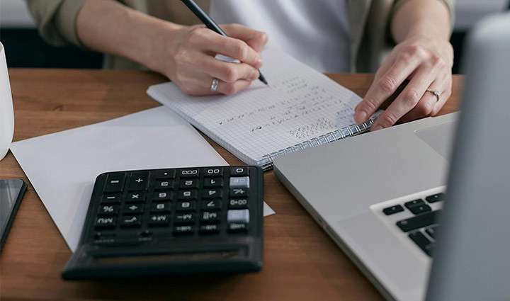 A warrior, wearing a tan jacket and white shirt, sits at a desk, writing notes on a notepad with a calculator and a laptop.