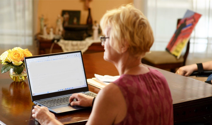 Christine Schei, an older woman with short blond hair, sits at her kitchen table typing on her laptop.