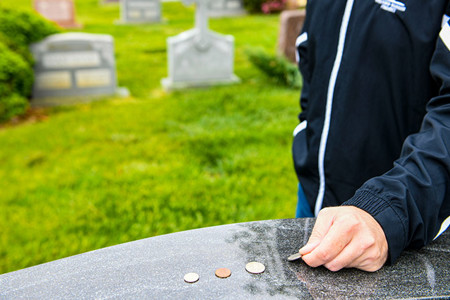 A person in a black jacket places a coin on a dark, polished gravestone with other coins present. Blurred white headstones are in the background.