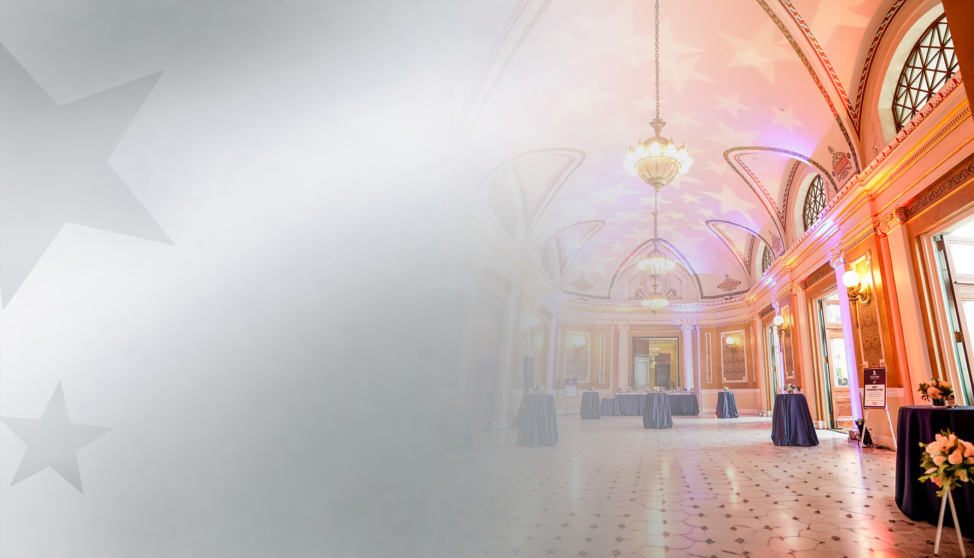 Elegant event hall with high arched ceilings, ornate chandeliers, and warm lighting, set with cocktail tables.