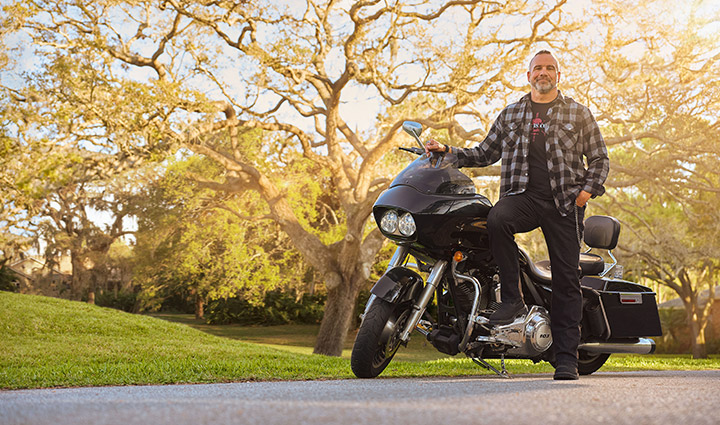Wounded warrior Bill Geiger stands alongside his motorcycle under a giant oak tree while smiling at the camera.