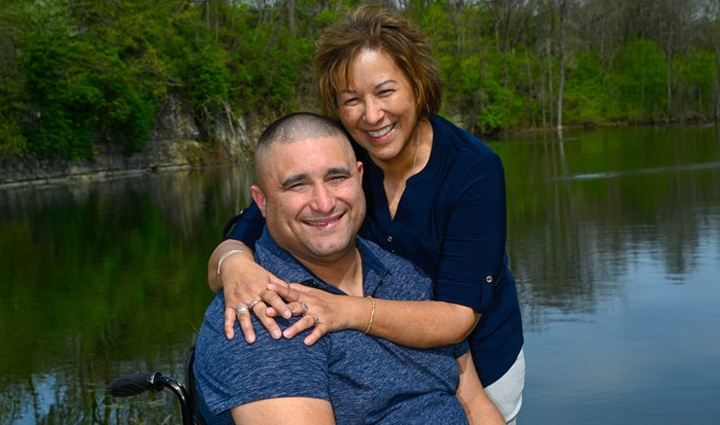 Caregiver Cindy Parsons smiles while hugging her adult son, Shane, seated in his wheelchair, also smiling. 