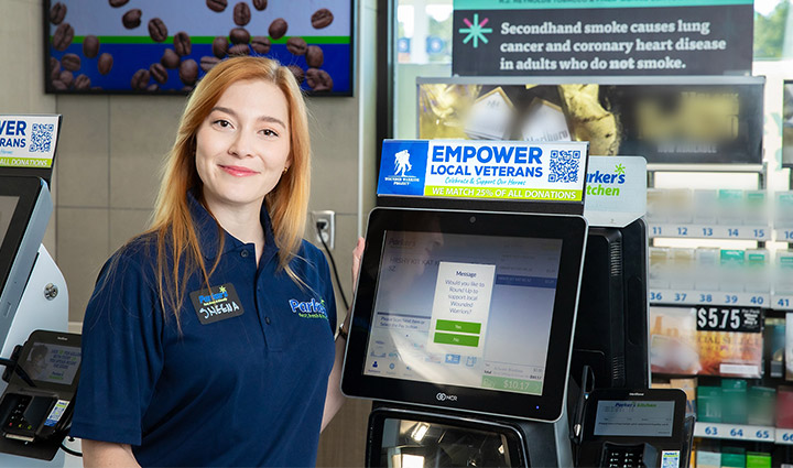 A brunette woman smiles while standing next to a check out computer screen in a convenience store.