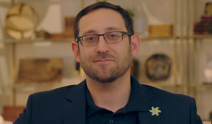A dark-haired man in a blue jacket sits doing an interview close to an out of sight camera.