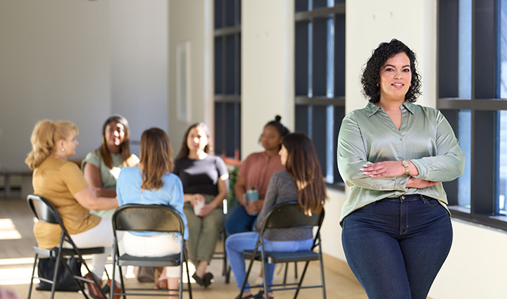 Wounded warrior Yomari Cruz stands near a group of women warriors seated in a circle talking at work.