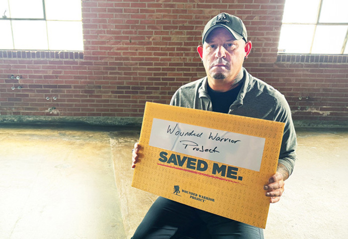 Warrior Phil Krabbe sits in front of a brick wall, holding a yellow sign that says, 