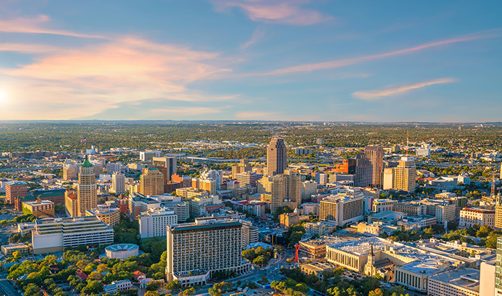 Aerial view of downtown San Antonio with Tower Life Building, Weston Centre, and sunlit skyline under a clear, cloud-dotted sky. 