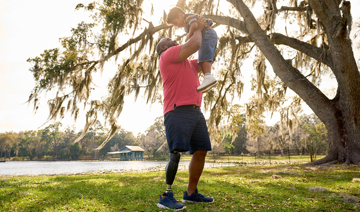 Wounded warrior Chris Gordon smiles while holding his son up in the air. Chris has a right prosthetic leg and wears a red t-shirt and blue shorts.
