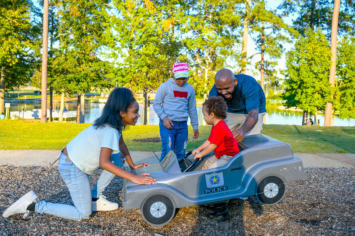 Wounded warrior Eric Delion and his oldest daughter play with his toddler as he sits in a child-sized police car. His other daughter looks on as they play.