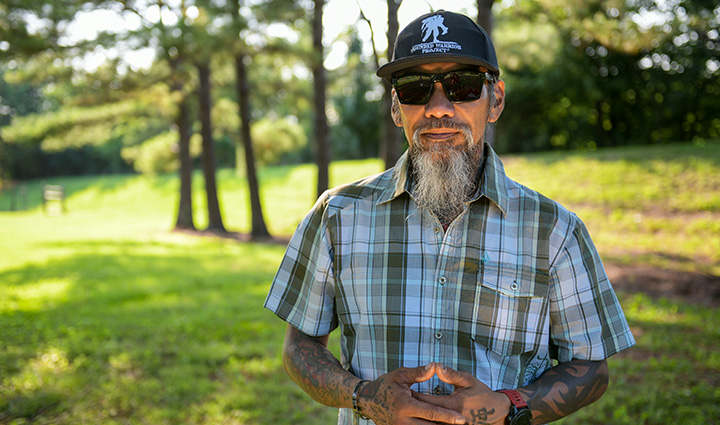 Wounded warrior Ray Andalio stands outside wearing sunglasses; trees in the background as he shows a slight smile to the camera. 