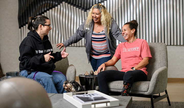 Warriors Beth King, Ysabel Cardona and Kelly Elmlinger gather in a lounge area, talking and seated in upholstered chairs near a small table.