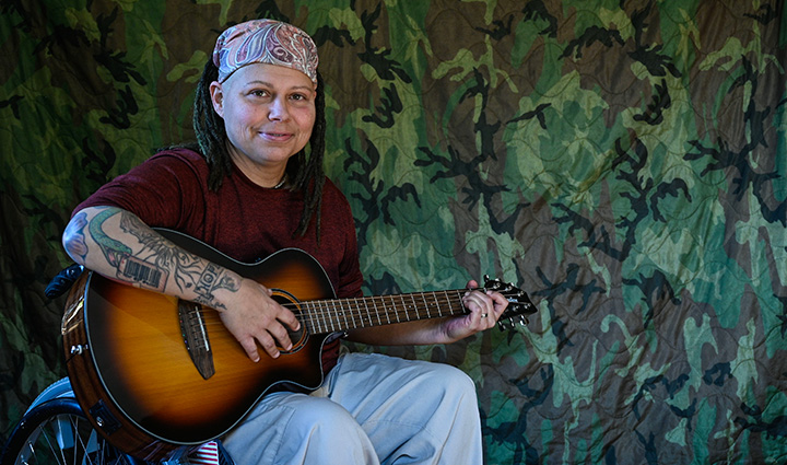 Wounded warrior Angie Lupe sits in her wheelchair in front of a camouflage background while holding her guitar. 
