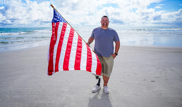 Wounded warrior Bryan Wagner, a single leg amputee, stands on the beach holding an American flag.