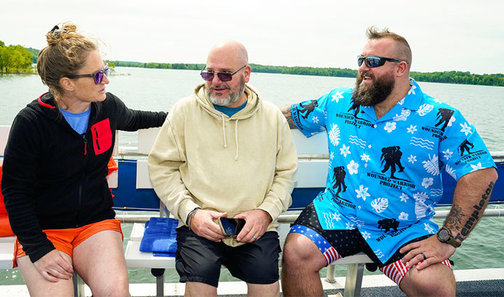 Warriors Elaine Schuett, Robert Kissee, and Matthew Templeton wearing sunglasses sit on a boat bench on a lake, talking together, with calm water and a tree-lined shoreline in the background.