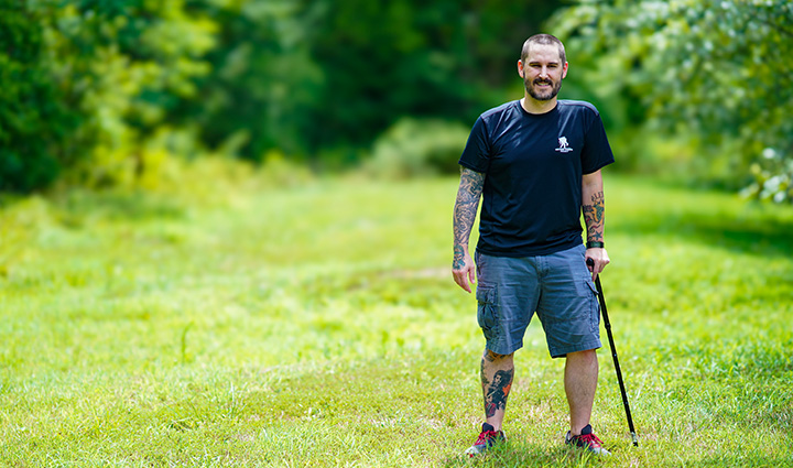 Wounded warrior Xander Hernandez stands in a field, smiling, while supporting himself with the use of a cane.